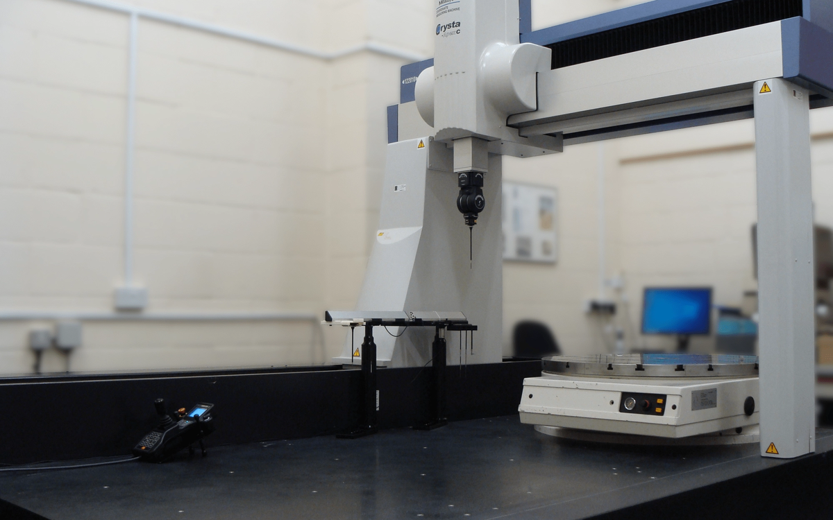 Five-axis scanning coordinate measuring machine with a vertical measurement head and rotary table in an inspection room.