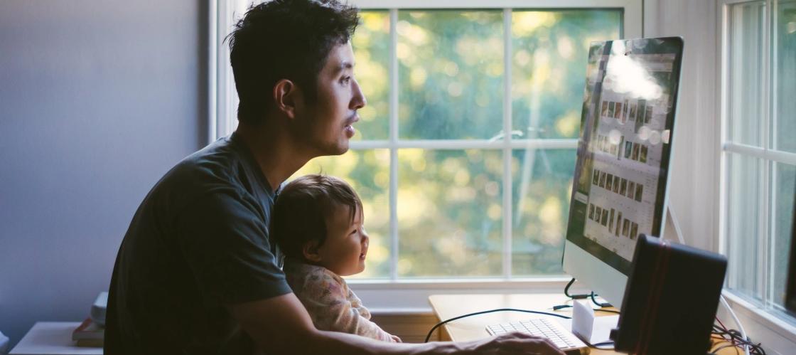 A man sitting in front of a computer screen, possibly mortgage shopping, while holding a baby in his lap