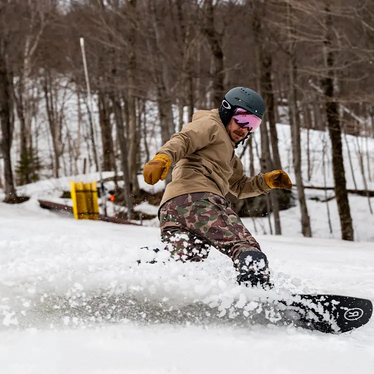snowboarder slashing a turn to the left while wearing camo snow pants and a tan winter coat