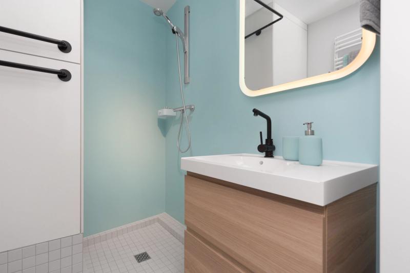 Prysznic otwarty, bez brodzika. Turkusowa ściana, lustro z podświetleniem, czarna bateria. Szafka wielofunkcyjna z pralko-suszarką — ta biała zabudowa po lewej.