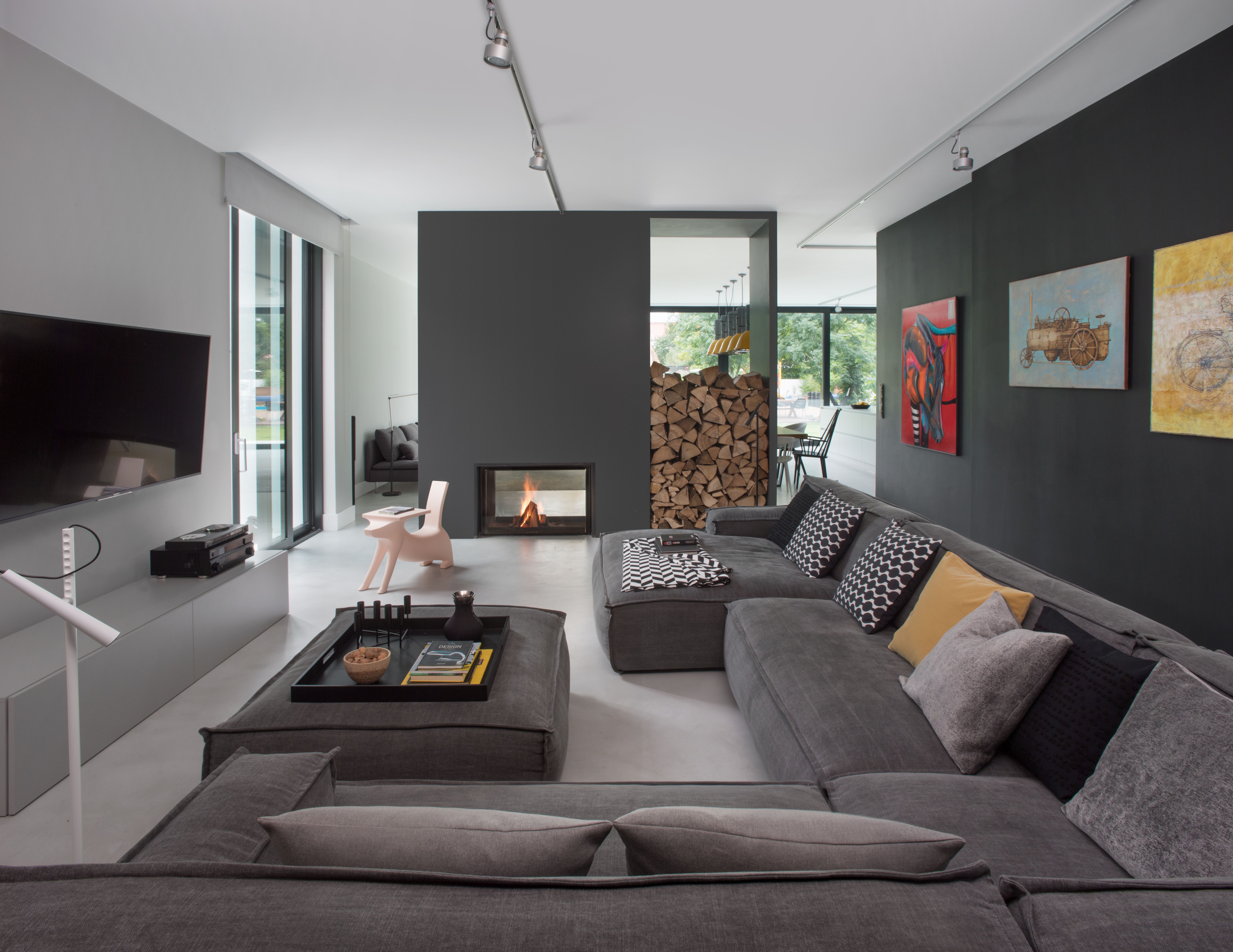 Nowoczesny pokój dzienny z szarą narożną sofą, czarną ścianą TV, drewnianym paleniskiem i sufitowym oświetleniem torowym