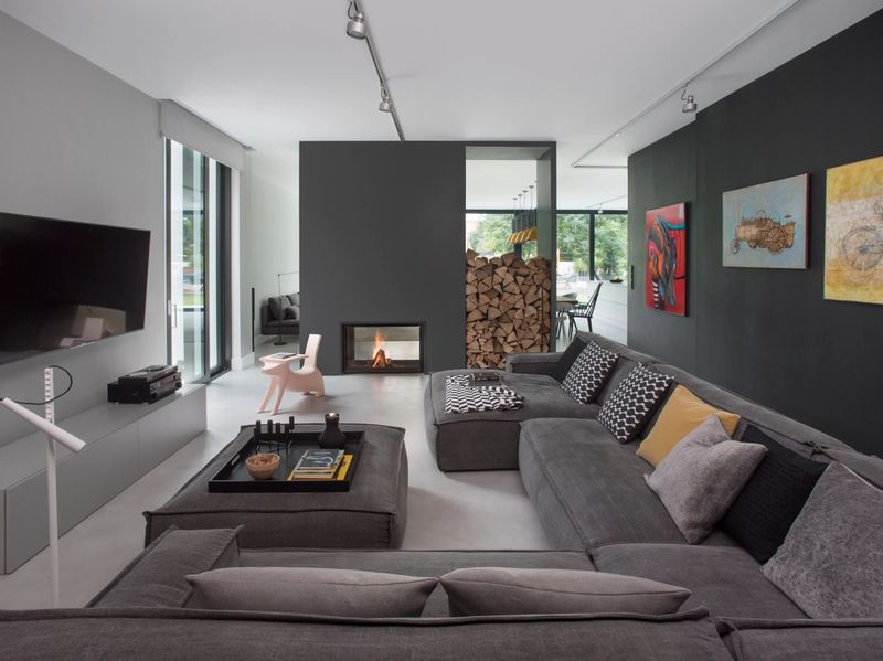 Nowoczesny pokój dzienny z szarą narożną sofą, czarną ścianą TV, drewnianym paleniskiem i sufitowym oświetleniem torowym