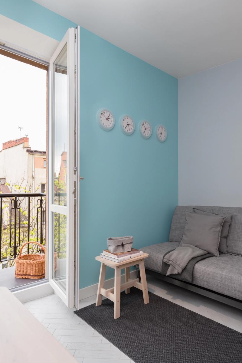 Kraków, Wellington, Berlin — zegary ze strefami czasowymi na turkusowej ścianie. Sofa rozkładana — śpią na niej kolejne dwie osoby.