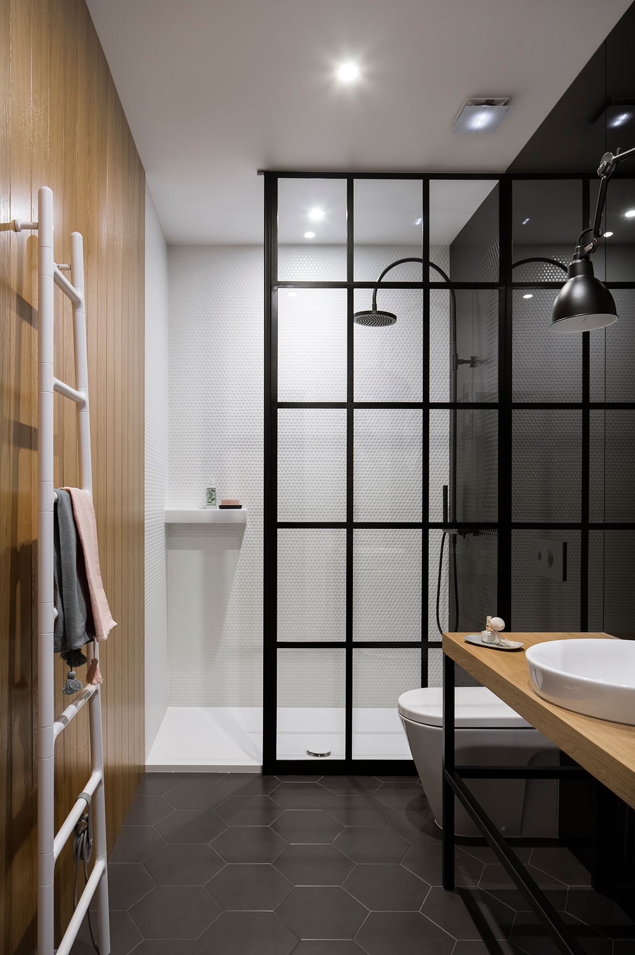 Nowoczesna łazienka z drewnianą ścianą, walk-in prysznicem za czarną ścianką oraz umywalką na drewnianym blacie. Białe płytki hexagonalne i czarne kafelki, elegancka armatura