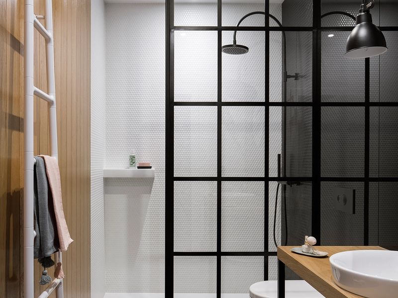 Nowoczesna łazienka z drewnianą ścianą, walk-in prysznicem za czarną ścianką oraz umywalką na drewnianym blacie. Białe płytki hexagonalne i czarne kafelki, elegancka armatura
