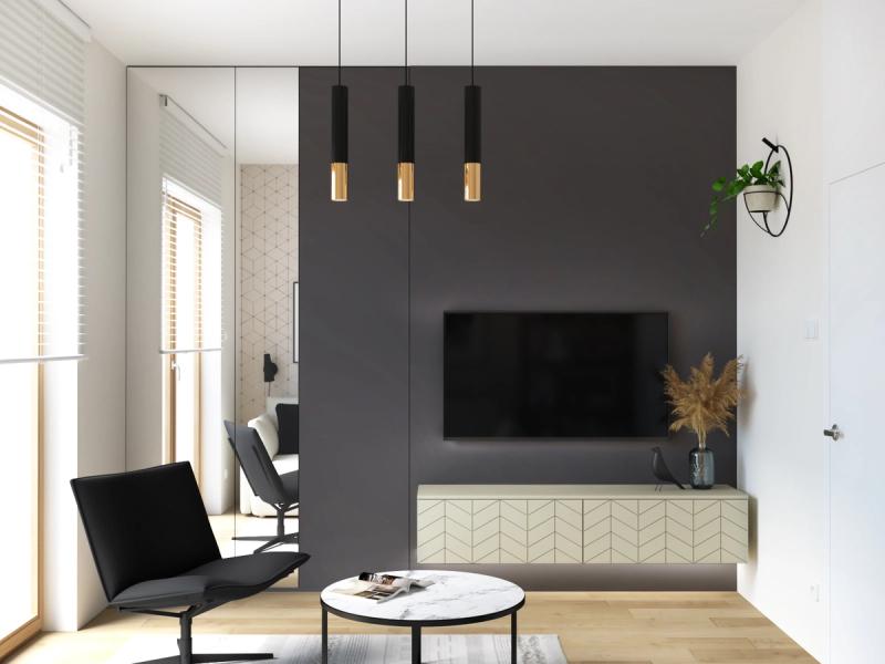 Grafitowa ściana — telewizor na niej nie dominuje. Trzy lampy: czarne tuby, mosiężne zakończenia. Szafka RTV z frontem w jodełkę. Fotel na metalowej nodze — jedyny mocny czarny akcent.