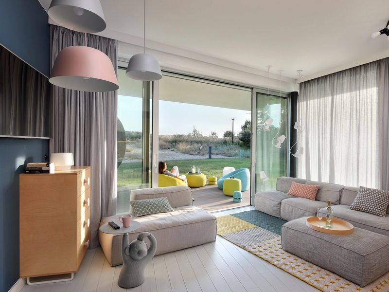 Nowoczesny salon z narożną sofą, ścianą TV w kolorze granatowym, regałem drewnianym i sufitowym oświetleniem. Panoramiczne okna, naturalne światło, minimalistyczne wyposażenie.