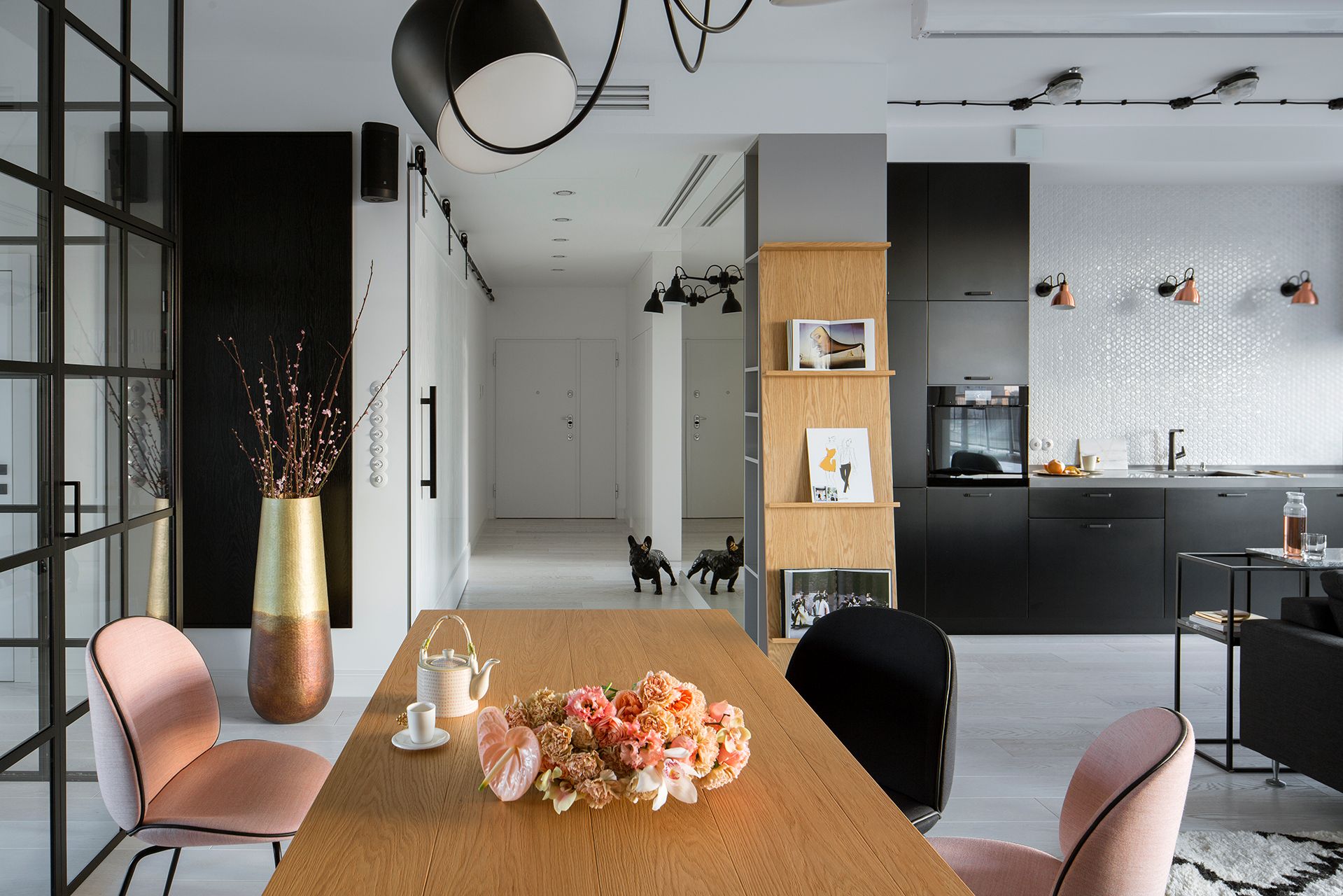 Nowoczesny salon z drewnianym stołem, różowymi krzesłami, regałem z drewna dębowego, czarną kuchnią i sufitowym oświetleniem w stylu minimalistycznym