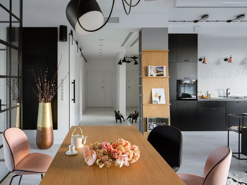 Nowoczesny salon z drewnianym stołem, różowymi krzesłami, regałem z drewna dębowego, czarną kuchnią i sufitowym oświetleniem w stylu minimalistycznym