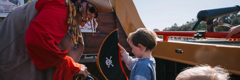 A crew member in a pirate hat and red coat hands skull-and-crossbones flags to two small children on deck