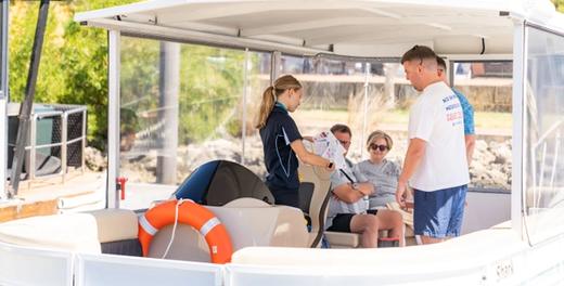 A Blue Manna crew member running through the safety briefing with three guests aboard a moored pontoon, an orange life ring in the foreground