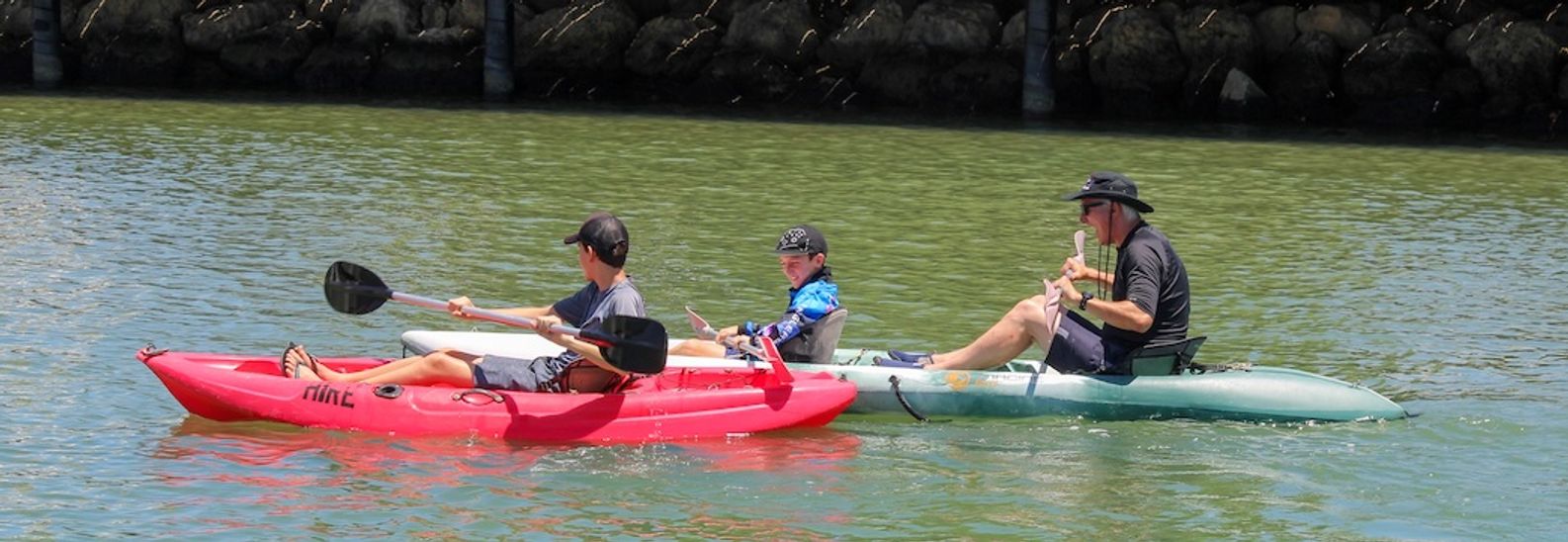 Adults and child paddling red and green kayaks past the Mandurah Boat Hire kiosk on a sunny day
