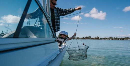 A man and a small boy in a captain's hat lowering a crab net over the side of a boat into the estuary