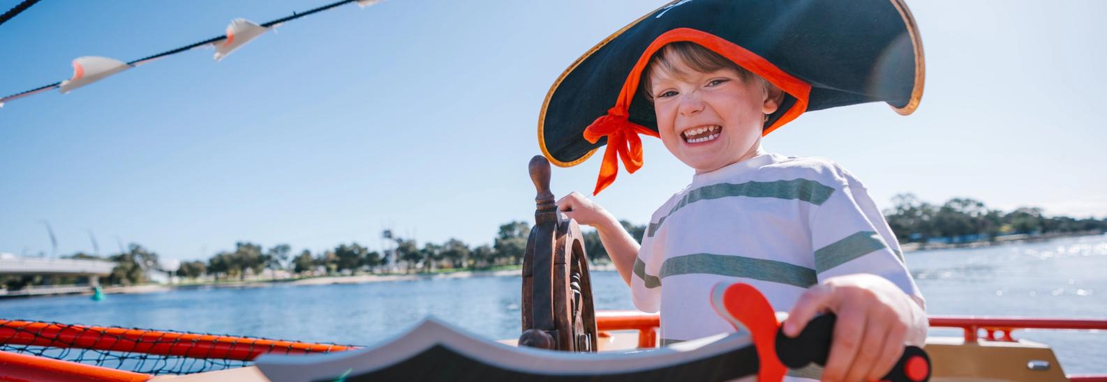 A grinning child in an oversized pirate hat holds a foam cutlass at the ship's wheel, the Mandurah estuary behind them