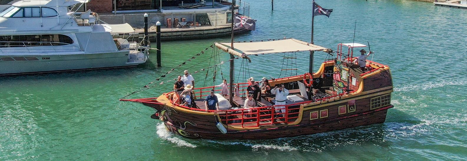 The pirate ship passing moored boats in the Mandurah canals, a costumed crew member on the stern deck