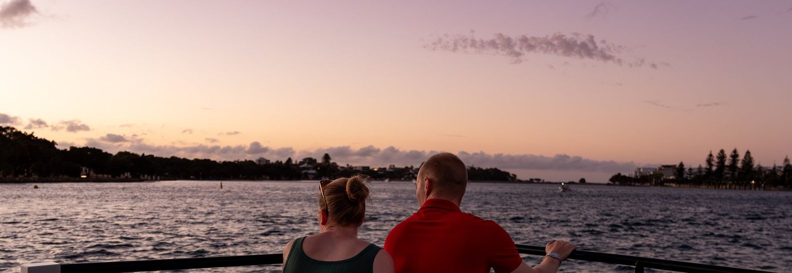 Couple leaning on black bow rails of a charter vessel watching the estuary at sunset