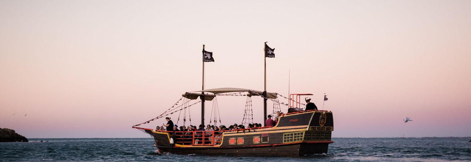 Pirate ship carrying seated guests under Jolly Roger flags on open water at dusk