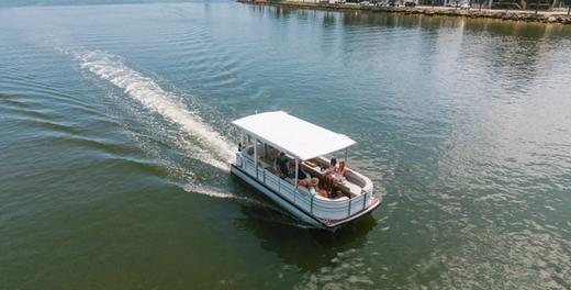 A hire pontoon moving through the water from above, with passengers on board and Mandurah's waterfront in the background