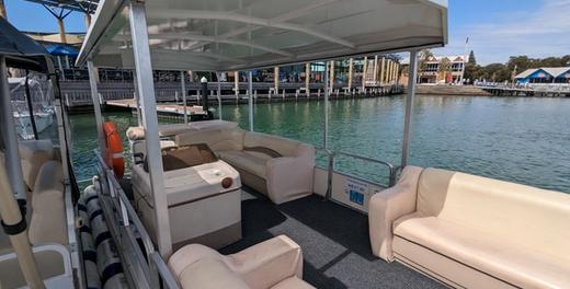 The deck of a hire pontoon, with cushioned bench seating under a white canopy, moored at the marina