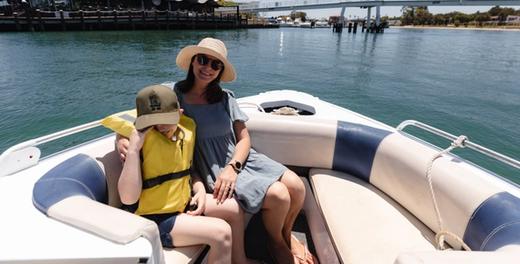 A woman and a child in a life jacket sitting in the bow of a hire boat, with the Mandurah footbridge and waterfront behind them