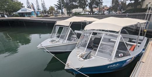Two runabouts with canvas canopies moored side by side at the jetty, with the Mandurah Boat Hire office behind them