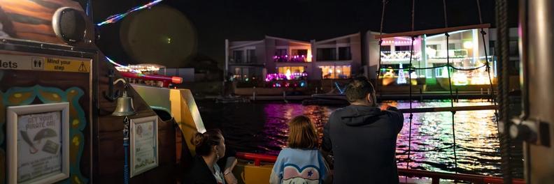 A family on the Pirate Ship's deck under strings of lights and skull-and-crossbones bunting, watching lit canal homes go past