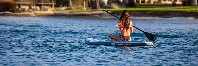Woman kneeling on an inflatable stand-up paddleboard, paddling estuary water near a road bridge