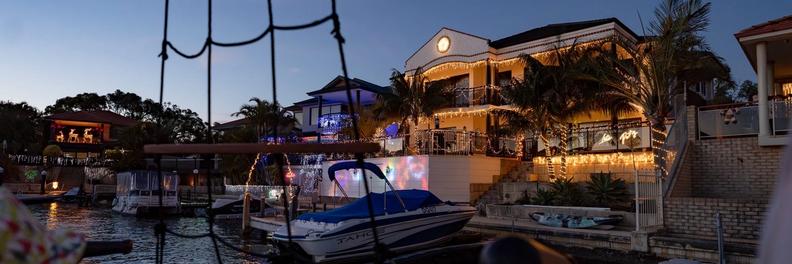 A canal home strung with warm white lights, seen through the ship's rigging while there is still blue in the sky