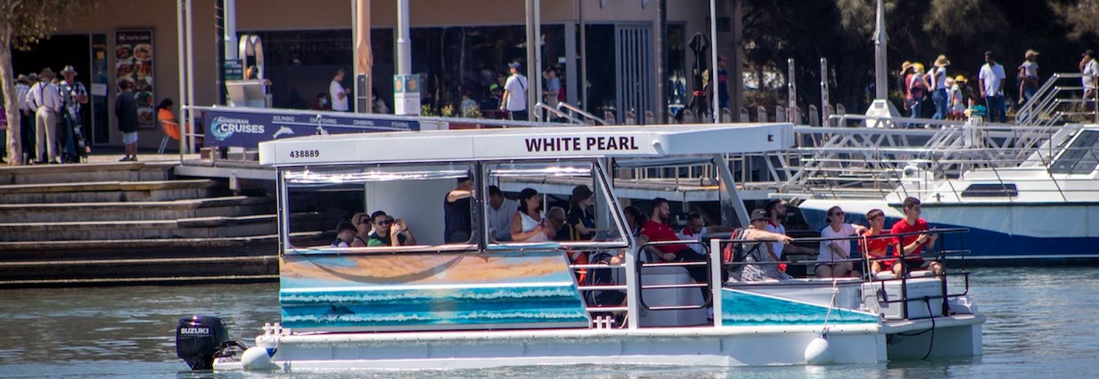 The White Pearl, a white pontoon boat with an open upper deck, cruising past the Mandurah waterfront with passengers aboard