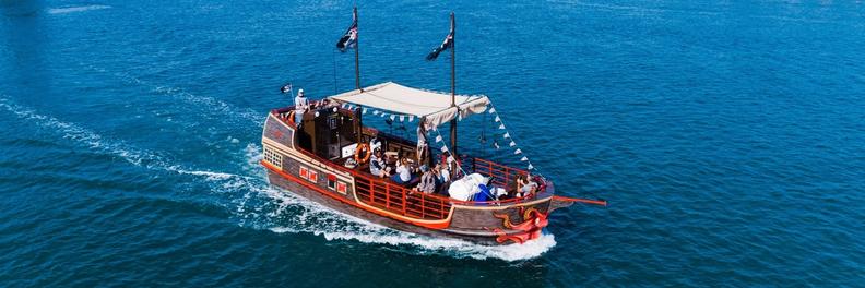The pirate ship under way on open blue water, pirate flag flying and passengers under the canopy