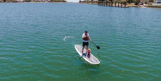 An adult paddling a stand-up paddleboard across the estuary with a small child kneeling on the front of the board