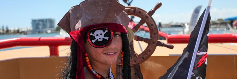 A child in full pirate costume with an eyepatch and dark wig stands beside the ship's wheel holding a flag