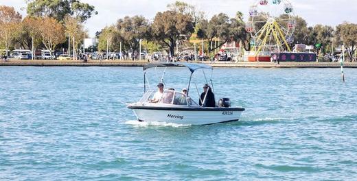 A small hire dinghy under way on the estuary with three people aboard, the foreshore and its Ferris wheel behind