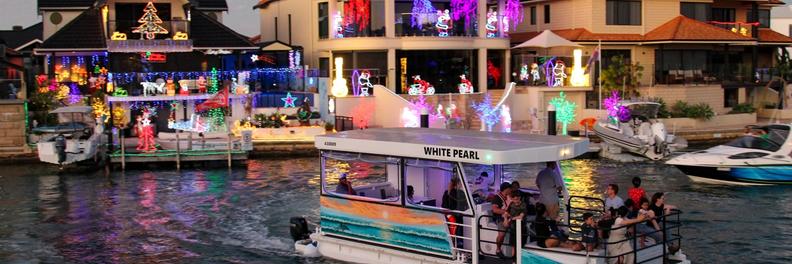 The White Pearl passing a row of canal homes covered in Christmas lights, passengers watching from the open rear deck