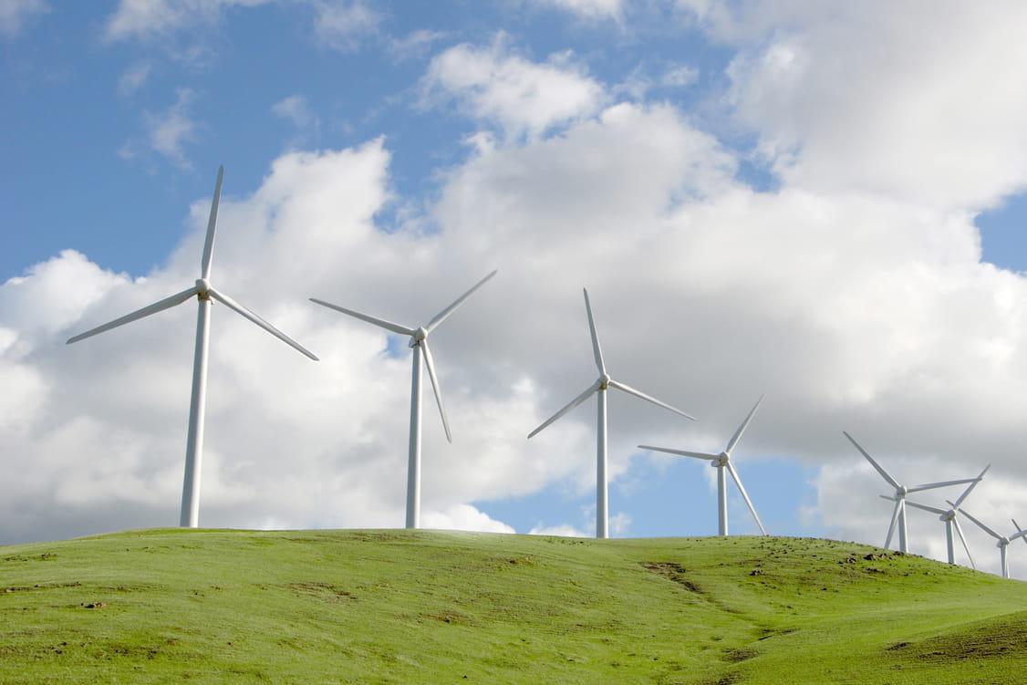 Wind Turbines seen from the foot of a hill covered in green grass with a sunny blue sky and clouds.