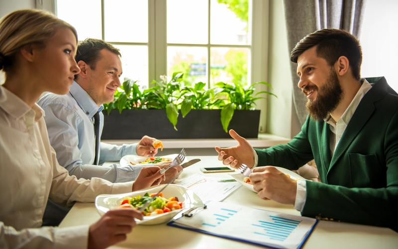 Three people in conversation over lunch at a sunlit table, with printed charts and papers beside them