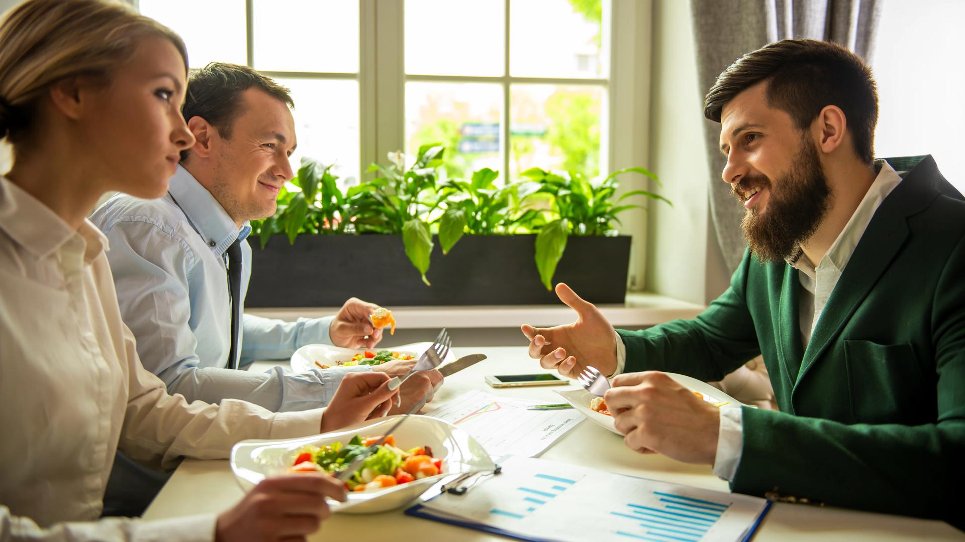Three people in conversation over lunch at a sunlit table, with printed charts and papers beside them