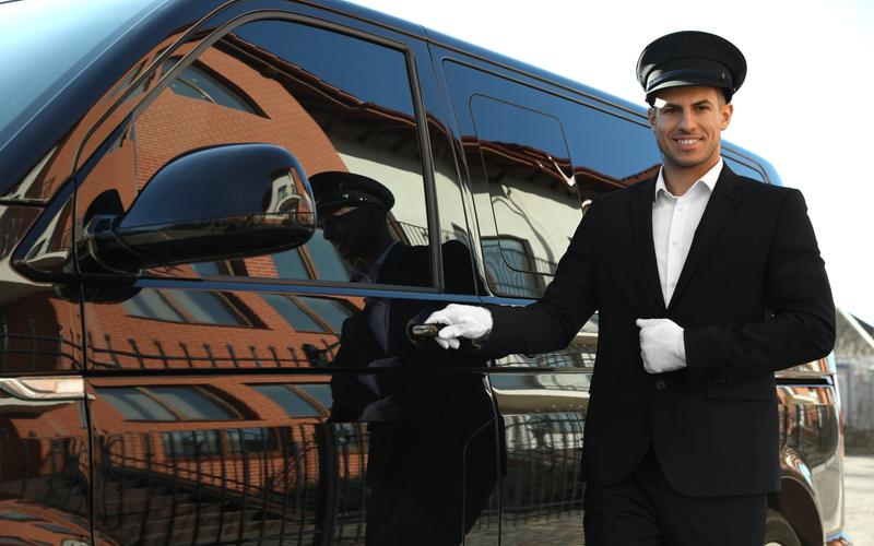 A chauffeur in a peaked cap and white gloves holding open the door of a black luxury car