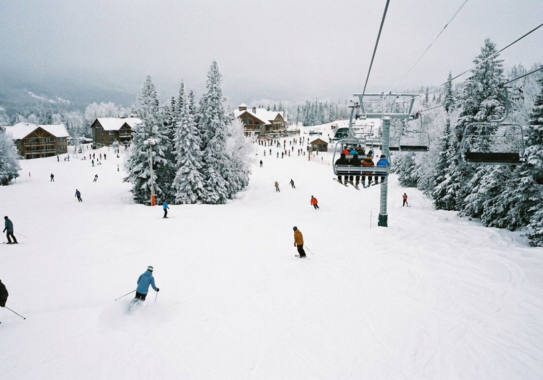 Skiers and snowboarders carve down wide, snow-covered runs at a North American ski resort in mid-winter, with a chairlift carrying people uphill and snow-laden evergreen trees along the slopes.