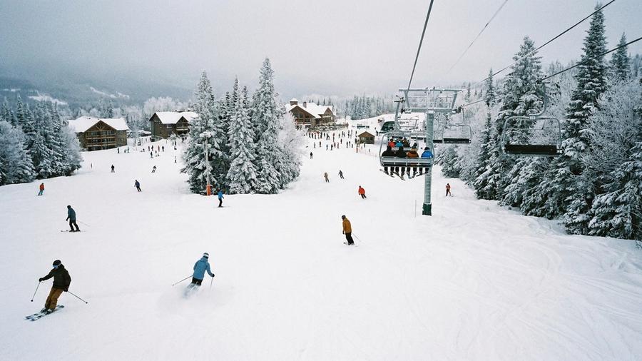 Skiers and snowboarders carve down wide, snow-covered runs at a North American ski resort in mid-winter, with a chairlift carrying people uphill and snow-laden evergreen trees along the slopes.