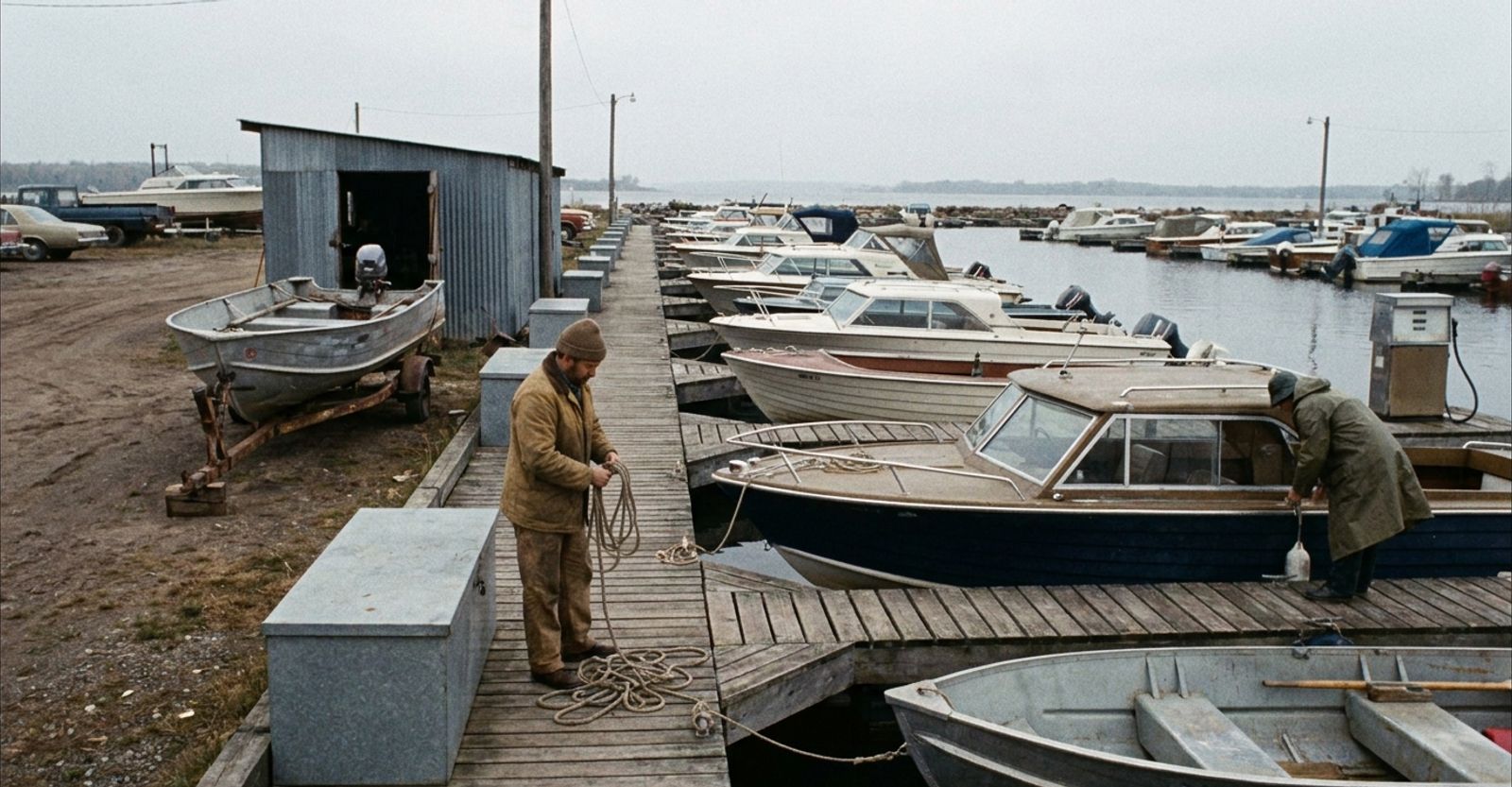 Overcast morning at a small working marina, with modest powerboats along a wooden dock, an aluminum fishing boat on a trailer beside a service shed, a fuel pump on the dock and two staff coiling lines and checking boats