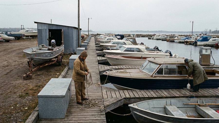 Overcast morning at a small working marina, with modest powerboats along a wooden dock, an aluminum fishing boat on a trailer beside a service shed, a fuel pump on the dock and two staff coiling lines and checking boats