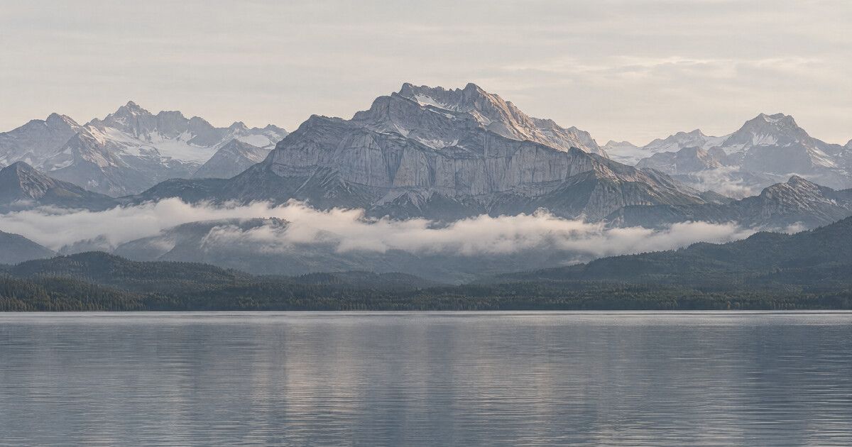 Alpenmassiv über einem Band tiefer Wolken, mit bewaldeten Hängen und einem ruhigen See im Vordergrund