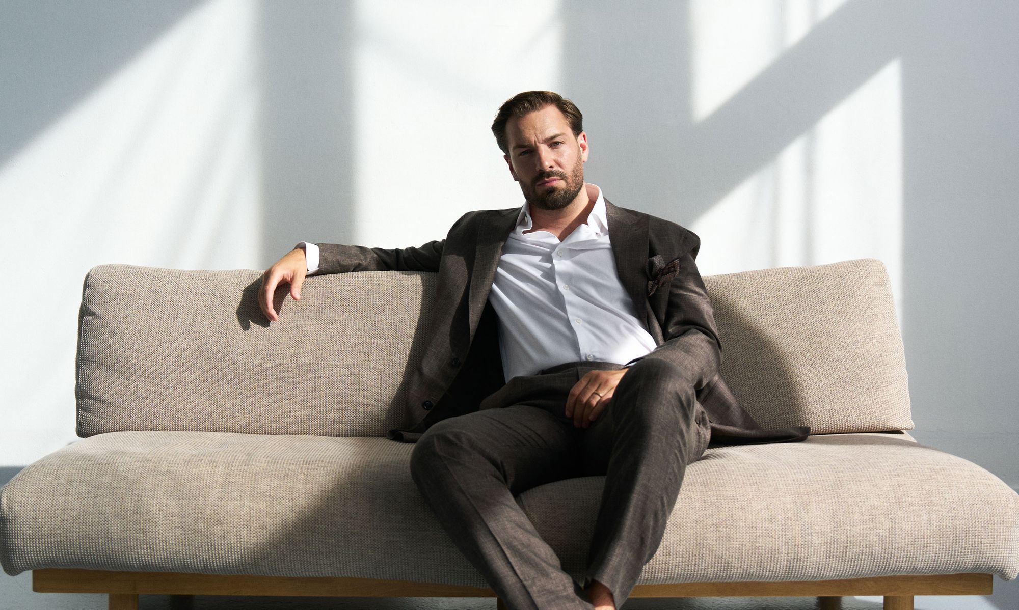 Portrait of Thibault Leroy Bürki in a dark suit and white shirt, seated on a beige sofa in natural light