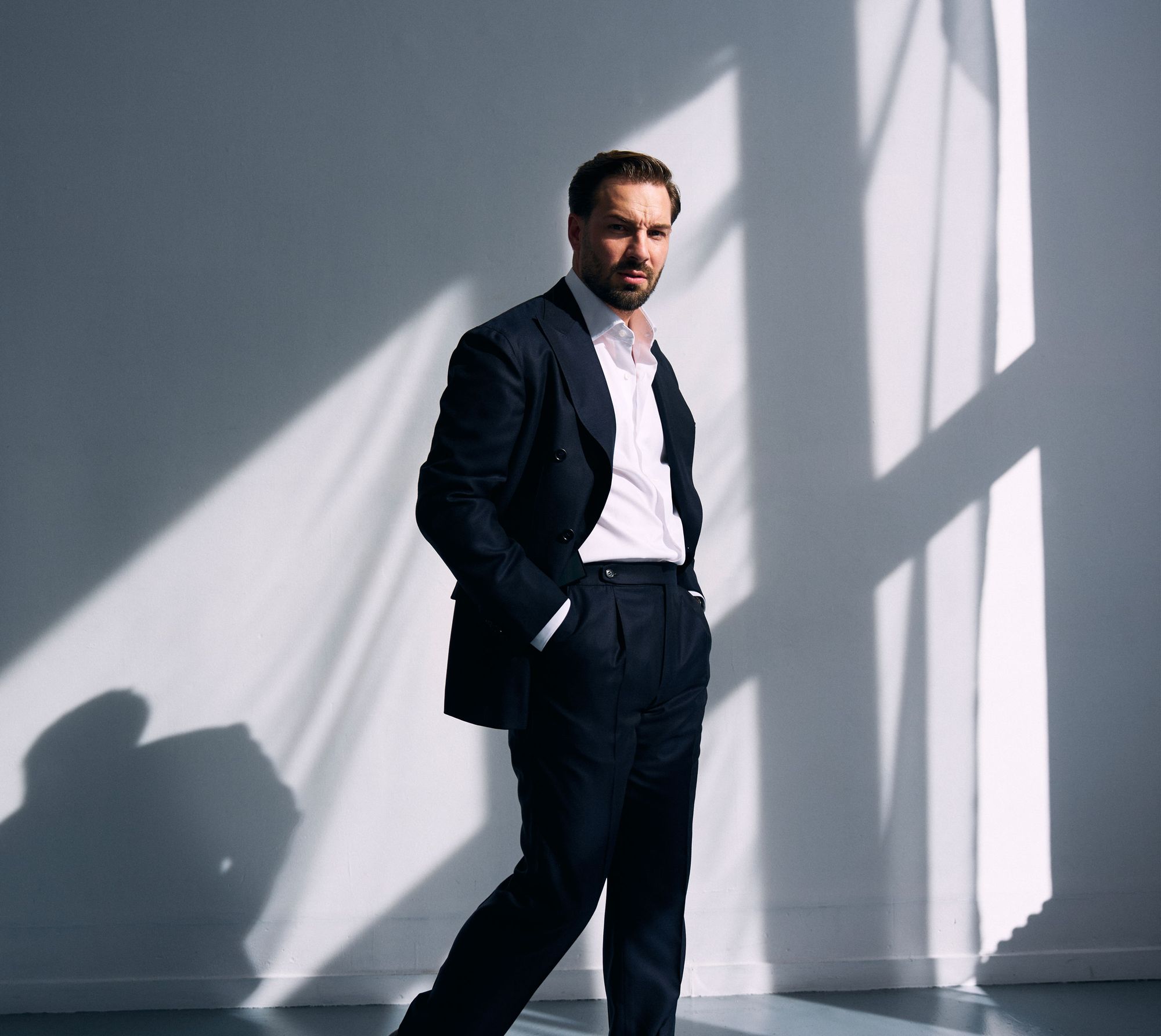 Portrait of Thibault Leroy Bürki standing in a dark suit and white shirt, hands in his pockets, against a sunlit wall