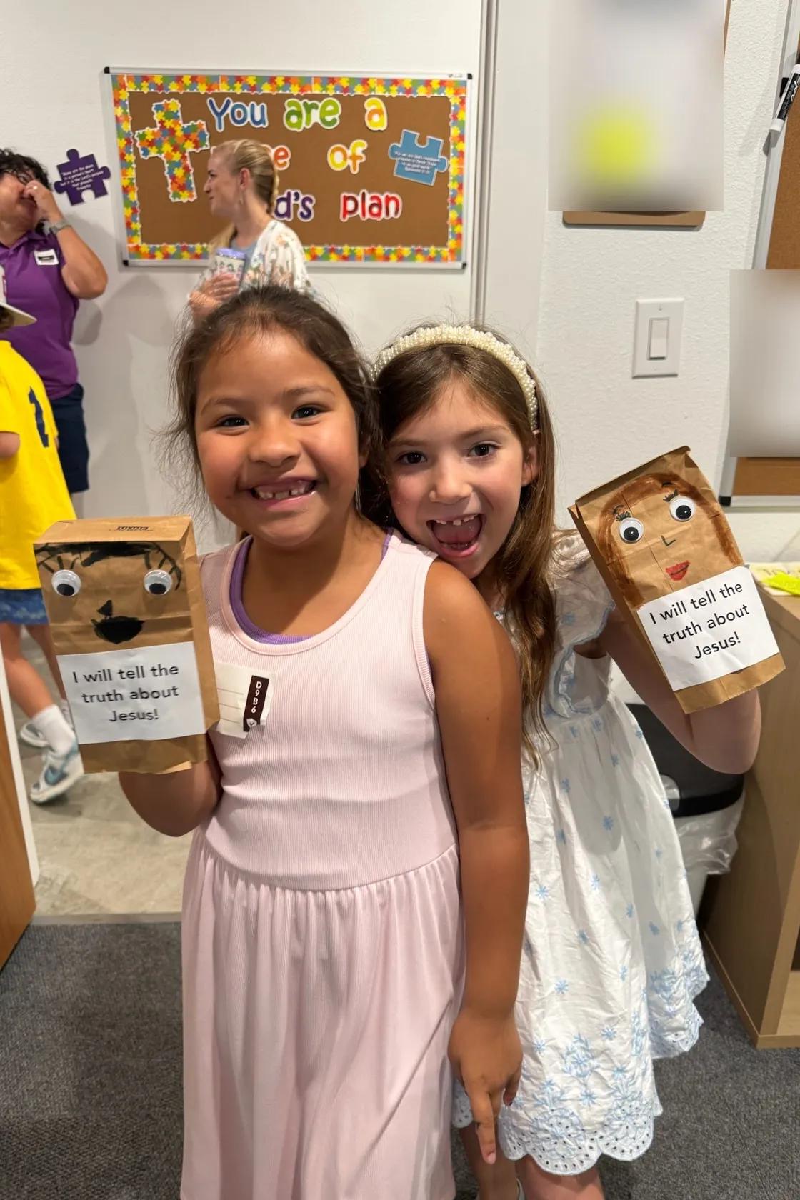 Two girls smiling with paper-bag puppets that read “I will tell the truth about Jesus!”