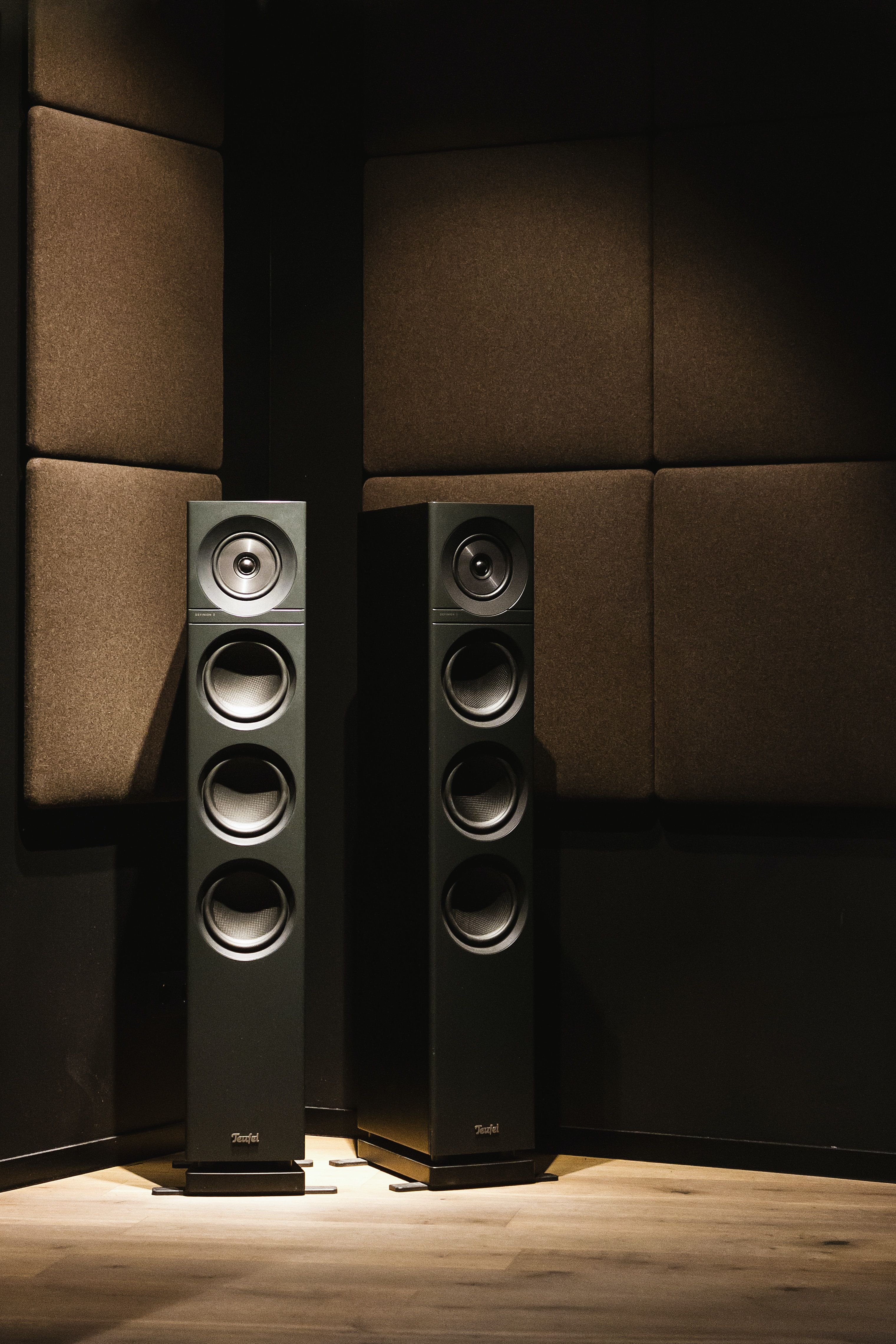 A pair of tall slim black speakers in the corner of a studio with padded walls