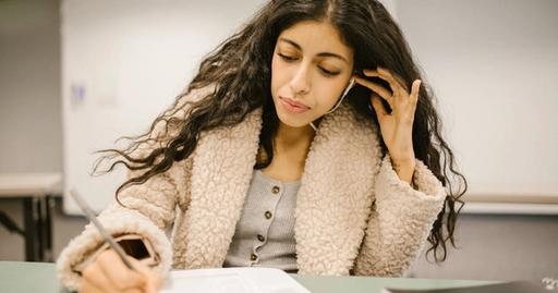 Female student writing in exam, using earphones for better focus in an educational setting.