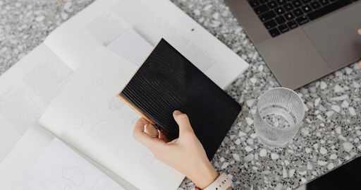 Overhead view of a hand holding a black notebook on a terrazzo table with a laptop and glass of water.