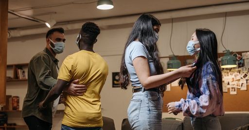 Friends gather indoors wearing masks, engaging in conversation amidst modern decor.